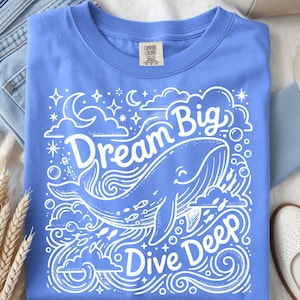 May include: A periwinkle blue t-shirt featuring a white graphic print of a whale swimming among clouds, stars, and bubbles. The text "Dream Big" is arched above the whale, and "Dive Deep" is printed below, all in a stylized, bold white font.