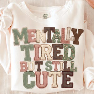 Può includere: Felpa color crema con la scritta "MENTALLY TIRED BUT STILL CUTE" in lettere colorate cucite. Le lettere sono in tonalità di verde, marrone, rosa e crema, alcune con motivi floreali. La felpa ha maniche lunghe.