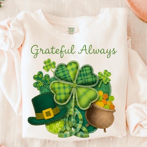 Puede incluir: Camiseta de manga larga color crema con las palabras "Grateful Always" en escritura verde. La camiseta presenta un gran diseño de trébol verde, un sombrero de duende verde y una olla de oro, perfecta para el Día de San Patricio.