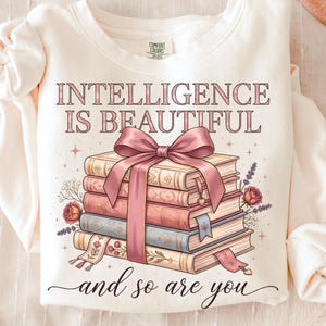 Puede incluir: Sudadera color crema con el texto "INTELLIGENCE IS BEAUTIFUL and so are you." El diseño presenta una pila de libros atados con un lazo rosa. Acentos florales y estrellas rodean los libros, creando una estética acogedora.
