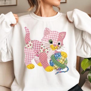 May include: White sweatshirt featuring a patchwork kitten design. The kitten is pink with floral, gingham, and polka dot patterns, holding a ball of yarn. The yarn ball has blue, green, and yellow accents. The kitten has yellow feet and a yellow mouth.