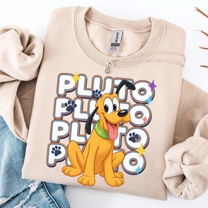 Op de afbeelding: Beige sweatshirt met een cartoon illustratie van Pluto, de Disney hond. Het woord "PLUTO" is drie keer gestapeld in een retro lettertype, met pootafdrukken en ster accenten. De sweatshirt is op een wit oppervlak gevouwen.