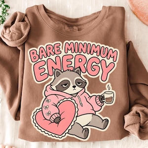 May include: A brown sweatshirt with the text "BARE MINIMUM ENERGY" in pink. The graphic features a raccoon wearing a pink hoodie and holding a coffee cup, with a heart-shaped pillow. The sweatshirt has long sleeves.