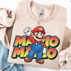 May include: Beige sweatshirt featuring a colorful graphic of Mario from the Super Mario Bros. video game. Mario is wearing his signature red cap, blue overalls, and white gloves. The text "MARIO" is in a vibrant, gradient font.