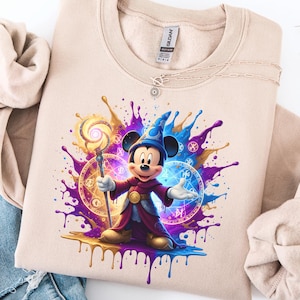 May include: Beige sweatshirt with a colourful graphic of Mickey Mouse dressed as a wizard, holding a staff. The design features splashes of purple, blue, and gold, with magical symbols. A necklace is visible near the collar.