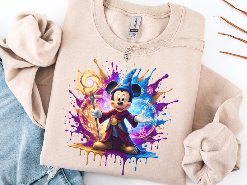 May include: Beige sweatshirt with a colourful graphic of Mickey Mouse dressed as a wizard, holding a staff. The design features splashes of purple, blue, and gold, with magical symbols. A necklace is visible near the collar.