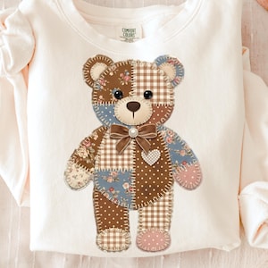 May include: Cream-colored sweatshirt featuring a patchwork teddy bear design. The bear has brown, blue, and floral patterned patches, a brown bow, and a heart-shaped detail. The sweatshirt has a Comfort Colors tag.