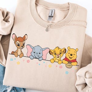 May include: Beige sweatshirt featuring a line of cartoon characters: Bambi, Dumbo, Simba, and Winnie the Pooh. The design includes colorful flowers and the word "Hunny" on a pot. A delicate necklace is draped across the top.