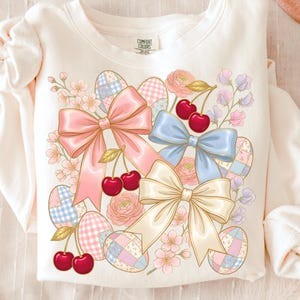 Könnte beinhalten: Cremefarbenes Sweatshirt mit einer farbenfrohen Grafik von Ostereiern, Schleifen, Kirschen und Blumen. Das Design umfasst rosa, blaue und cremefarbene Schleifen, mit roten Kirschen und pastellfarbenen Eiern. Der Text "Comfort Colors" ist oben sichtbar.