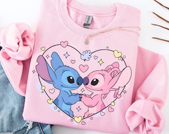 Könnte beinhalten: Hellrosa Sweatshirt mit herzförmigem Design mit den Zeichentrickfiguren Stitch und Angel. Die blauen und rosa Charaktere halten sich an den Händen, umgeben von Herzen und Sternen. Eine zarte Halskette mit Sonnenanhänger liegt auf dem Sweatshirt.
