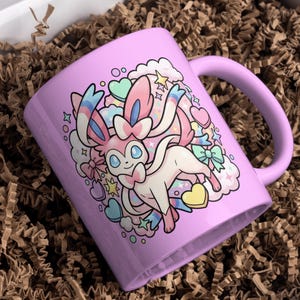 K&ouml;nnte beinhalten: Eine lavendelfarbene Keramiktasse mit einer Cartoon-Illustration eines Sylveon-Charakters. Das Design umfasst Herzen, Sterne und Wolken in Pastellfarben. Die Tasse hat einen gebogenen Henkel und befindet sich in einer Schachtel mit braunem Verpackungsmaterial.