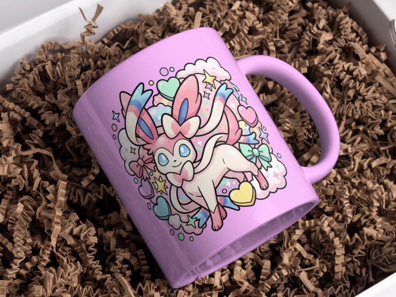K&ouml;nnte beinhalten: Eine lavendelfarbene Keramiktasse mit einer Cartoon-Illustration eines Sylveon-Charakters. Das Design umfasst Herzen, Sterne und Wolken in Pastellfarben. Die Tasse hat einen gebogenen Henkel und befindet sich in einer Schachtel mit braunem Verpackungsmaterial.