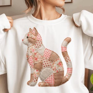 Op de afbeelding: Witte sweatshirt met een patchwork kattenontwerp. De kat is samengesteld uit verschillende stofpatronen, waaronder bloemen-, ruit- en hartprints, in tinten roze, beige en bruin. De kat zit met zijn staart gekruld.