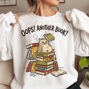 Op de afbeelding: Witte sweatshirt met de tekst "OOPS! ANOTHER BOOK!" boven een cartoon uil met een bril die op een stapel boeken zit. De boeken hebben verschillende kleuren en de uil ziet er moe uit. Een bril ligt ervoor.