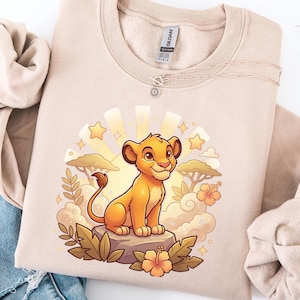 K&ouml;nnte beinhalten: Beiges Sweatshirt mit einer Cartoon-L&ouml;wenjungen-Illustration. Das Design zeigt ein l&auml;chelndes L&ouml;wenjunges, Sterne, Wolken und Blumen. Das Sweatshirt hat einen Rundhalsausschnitt und lange &Auml;rmel. Eine silberne Halskette mit Sonnenanh&auml;nger ist ebenfalls sichtbar.