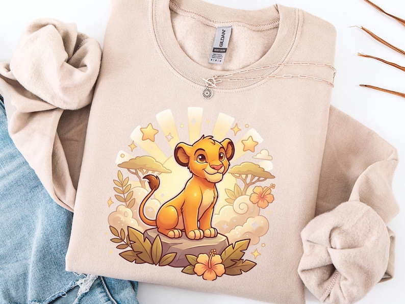 K&ouml;nnte beinhalten: Beiges Sweatshirt mit einer Cartoon-L&ouml;wenjungen-Illustration. Das Design zeigt ein l&auml;chelndes L&ouml;wenjunges, Sterne, Wolken und Blumen. Das Sweatshirt hat einen Rundhalsausschnitt und lange &Auml;rmel. Eine silberne Halskette mit Sonnenanh&auml;nger ist ebenfalls sichtbar.