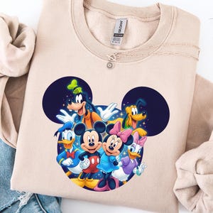 May include: A beige sweatshirt featuring a colorful graphic of Mickey Mouse, Minnie Mouse, Donald Duck, Daisy Duck, Goofy, and Pluto. The characters are arranged in a circle, with Mickey and Minnie in the center. A silver necklace with a sun pendant is on the sweatshirt.
