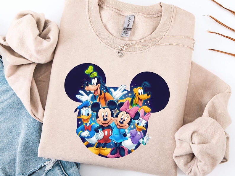 May include: A beige sweatshirt featuring a colorful graphic of Mickey Mouse, Minnie Mouse, Donald Duck, Daisy Duck, Goofy, and Pluto. The characters are arranged in a circle, with Mickey and Minnie in the center. A silver necklace with a sun pendant is on the sweatshirt.