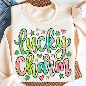 May include: Cream-colored sweatshirt with the words "Lucky Charm" in a colorful, whimsical design. The text is outlined in black and filled with green, blue, pink, and yellow hues, with shamrocks, hearts, and stars. The sweatshirt is folded on top of blue jeans.