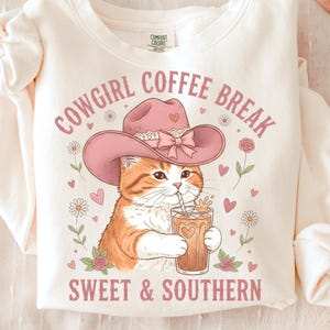 Può includere: Felpa color crema con grafica di un gatto con cappello da cowboy rosa che beve caffè freddo. Il testo recita "Cowgirl Coffee Break Sweet & Southern". Il design include cuori, fiori e un fiocco.