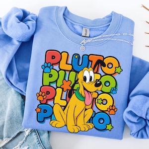 May include: A light blue sweatshirt featuring a colorful graphic of Pluto the dog with the word "PLUTO" in large, playful letters. The design includes paw prints and stars. A silver necklace is draped across the shirt.