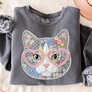 May include: Gray sweatshirt featuring a patchwork cat face with heart-shaped pink glasses and floral accents. The cat's face is made of various patterned fabrics, including floral and polka dot designs. The sweatshirt is a Comfort Colors brand.