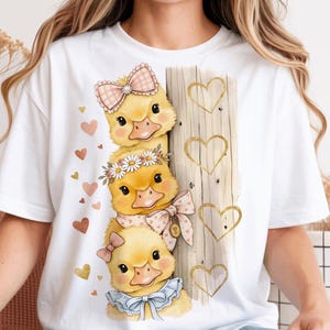 Może przedstawiać: Biała koszulka z ilustracją trzech żółtych kaczątek z kokardkami i kwiatowymi akcentami. Projekt zawiera drewnianą deskę z wycięciami w kształcie serca i rozrzuconymi sercami w odcieniach różu i złota.