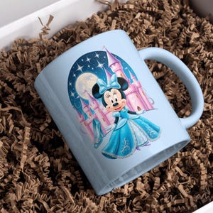 May include: A light blue ceramic mug with a colorful illustration of Mickey Mouse dressed as a princess in front of a castle. The background is a night sky with stars and a moon.