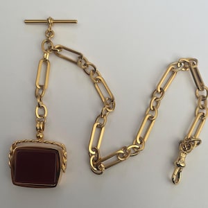 Op de afbeelding: Een goudkleurige zakhorlogeketting met een rechthoekige, roodbruine stenen hanger. De ketting heeft een T-bar, langwerpige schakels en een sluiting. De hanger is omlijst met een gedraaid touwontwerp.