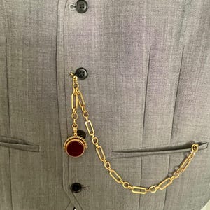 May include: A gold-colored pocket watch chain with a decorative fob and a red stone, attached to a gray vest. The chain is made of rectangular links and hangs from a button on the vest.