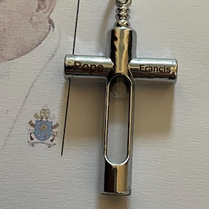 Puede incluir: Un colgante de cruz plateado con las palabras "Pope Francis" grabadas. El colgante tiene un lazo en la parte superior para una cadena y un centro hueco. El fondo incluye una foto de una persona y un escudo de armas.