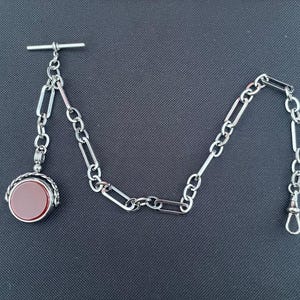May include: A silver-toned pocket watch chain with a T-bar, rectangular links, and a swivel clasp. The chain connects to a round pendant with a red-brown stone, possibly carnelian, set in a decorative bezel.