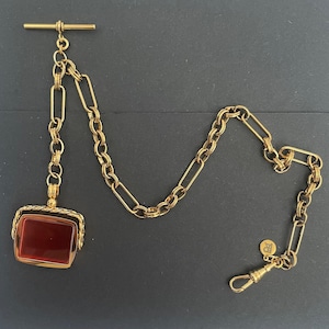 Puede incluir: Una cadena de reloj de bolsillo dorada con un colgante rectangular de piedra roja. La cadena presenta una barra en T, varios estilos de eslabones y un cierre. El colgante está enmarcado en oro y tiene una superficie pulida. La cadena está sobre fondo oscuro.