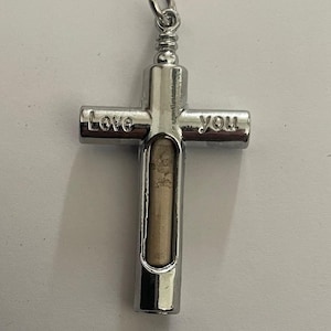 May include: A silver-colored cross pendant with the words "Love you" engraved on the arms. The cross has a cylindrical compartment containing a small, light-colored object. The pendant has a loop for attaching to a chain.