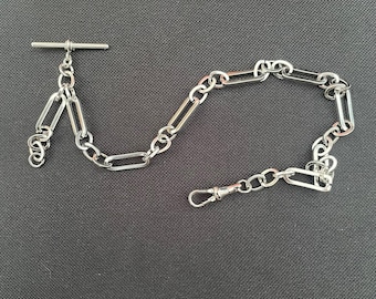 May include: A silver-colored watch chain with a T-bar and a clasp. The chain is composed of alternating oval and circular links. The T-bar is at the top, and the clasp is at the bottom. The chain is displayed on a dark gray surface.