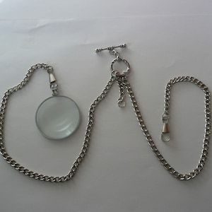 May include: A silver-toned monocle with a chain and a T-bar clasp. The monocle has a circular lens and is attached to a curb chain. The chain also has a spring-loaded clasp. The monocle is likely made of metal.