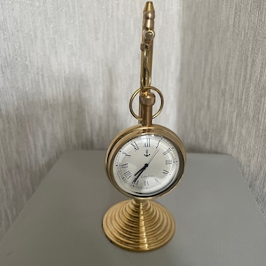 May include: A gold-colored desk clock with a round face and Roman numerals. The clock features an anchor symbol and the word "London" on the face. The clock is mounted on a decorative, spiral-shaped base.