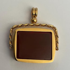 Può includere: Ciondolo dorato con un centro in pietra rettangolare rosso scuro. Il ciondolo ha un bordo dorato a forma di corda e un piccolo anello nella parte superiore per una catena. Il design è classico ed elegante, adatto per occasioni formali o quotidiane.