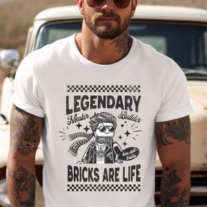 May include: White t-shirt featuring a black and white graphic of a Lego figure in sunglasses, a scarf, and a jacket. The text reads "LEGENDARY Master Builder BRICKS ARE LIFE" with a heart-shaped "love and bricks" graphic.