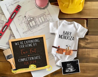 以下が含まれることがあります： 「We're expanding our home by two feet. Completion date: Feb. 2024」と書かれた黒い文字盤。白いロンパースには「Baby Mendoza」の文字と、ハンマー、ドライバー、ペンチがついたツールベルトが描かれている。画像には、黄色いヘルメット、赤いレベル、メジャー、ペンキ缶も写っている。