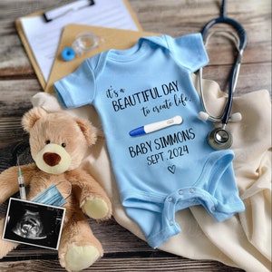 May include: A light blue baby onesie with the text "it's a beautiful day to create life."  A pregnancy test with a positive result is shown on the onesie. The text "Baby Simmons Sept. 2024" is also on the onesie.