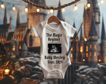 May include: White baby onesie with a black and white ultrasound image, the text "The Magic Begins!" and "Baby Wesley Sept. 2024" and a heart. The onesie is hanging from a rope against a blurry background of a castle.