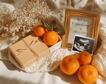 May include: A gold framed announcement with the text "We're Expecting! Mar. 2025" next to an ultrasound image. Oranges and a gift box tied with twine are arranged on a white crocheted blanket.