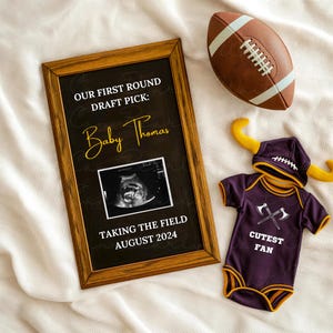 Op de afbeelding: Een ingelijst krijtbord-aankondiging met een zwarte achtergrond en witte tekst die luidt "Onze eerste ronde draft pick: Baby Thomas" met een echobeeld en de tekst "Het veld betreden augustus 2024". Een paars-gouden baby-romper met een voetbalhelm en de tekst "Schattigste fan" ligt op een witte deken met een bruine voetbal.