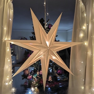 May include: A large, white paper star lantern hanging in a window with a Christmas tree decorated with purple and silver ornaments in the background.