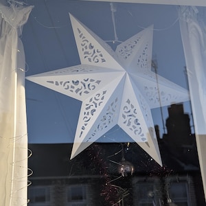 May include: A white paper star with a lacy design hangs in a window. The star is illuminated by the sun, casting a shadow on the wall behind it.