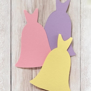 Pu&ograve; includere: Tre sagome di carta di abiti in diversi colori: rosa, viola e giallo. Gli abiti sono disposti a forma di triangolo su uno sfondo di legno chiaro.