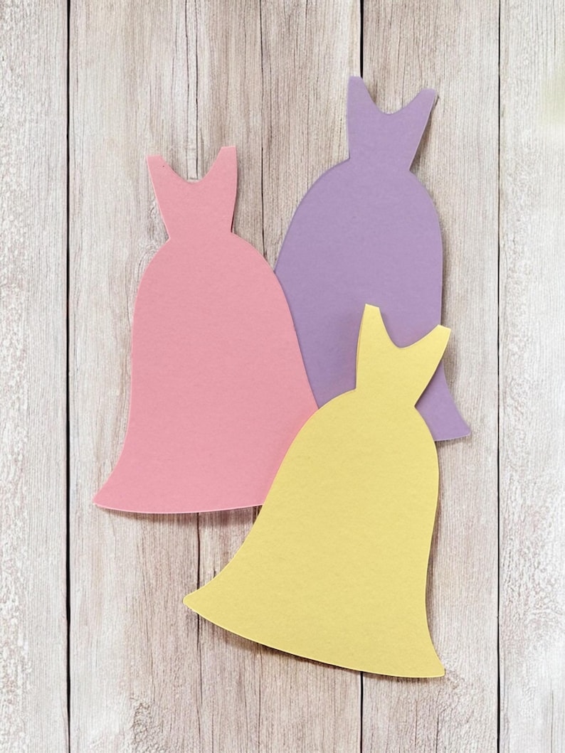Pu&ograve; includere: Tre sagome di carta di abiti in diversi colori: rosa, viola e giallo. Gli abiti sono disposti a forma di triangolo su uno sfondo di legno chiaro.