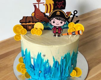 Può includere: Una torta a tema pirata blu e bianca con una figura di pirata dei cartoni animati, una nave e monete d'oro. La torta è decorata con glassa blu e codette argentate.