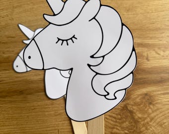 Puede incluir: Dos recortes de cabeza de unicornio blancos con contornos negros y ojos cerrados, unidos a palitos de madera. Los unicornios tienen un cuerno, una melena fluida y están sobre un fondo de madera.